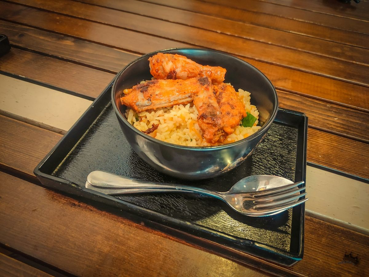 A bowl of chicken and rice on a tray with a fork and spoon, on a wooden table